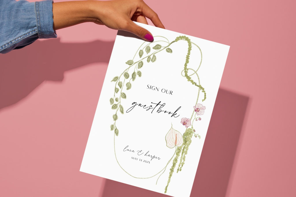 Floral wedding guestbook sign held against a pink background, featuring “please sign our guestbook” wording inside a curved green botanical border with burgundy flowers and romantic script lettering.