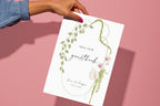 Floral wedding guestbook sign held against a pink background, featuring “please sign our guestbook” wording inside a curved green botanical border with burgundy flowers and romantic script lettering.