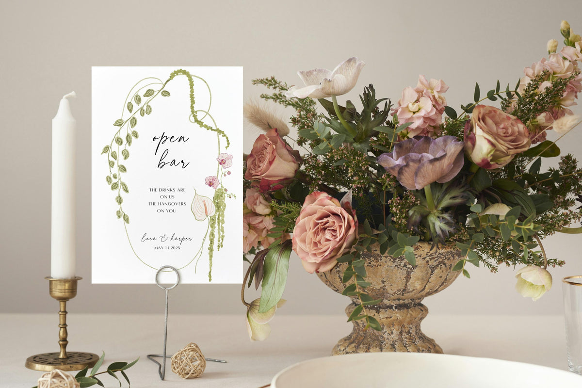Printable floral open bar wedding sign displayed beside a candle and flower arrangement, featuring a green botanical bow border for romantic garden wedding reception décor.