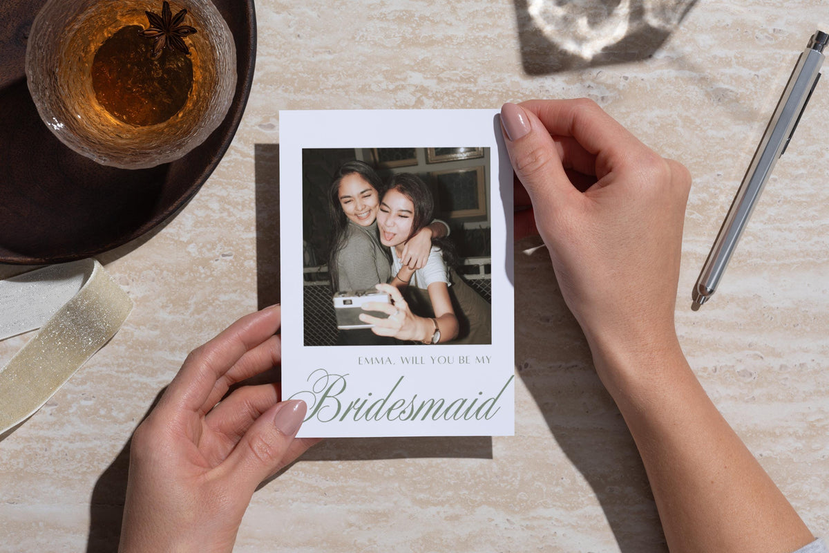 Hands holding a personalised photo bridesmaid proposal card above a stone table beside coffee and a pen. Sage green calligraphy and a friendship photograph create a sentimental will you be my bridesmaid keepsake.
