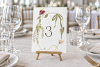 Editable botanical wedding table number template with hand-painted wildflowers, displayed on a wooden easel in an elegant reception setting.