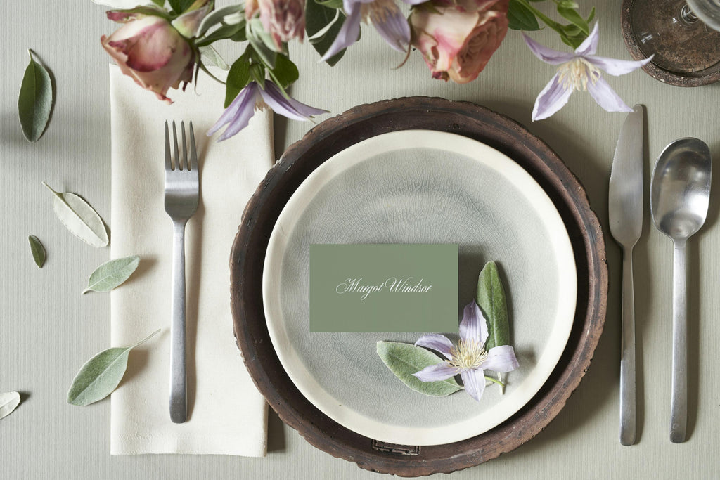 Sage green wedding place card with white calligraphy on stacked neutral ceramic plates, styled with silver cutlery and delicate flowers for a timeless garden wedding table setting.