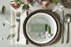 Sage green wedding place card with white calligraphy on stacked neutral ceramic plates, styled with silver cutlery and delicate flowers for a timeless garden wedding table setting.