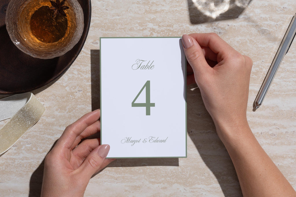 Sage green wedding table number template viewed from above, held over a textured neutral table beside a drink and cutlery, with refined serif typography for understated old money wedding reception décor.