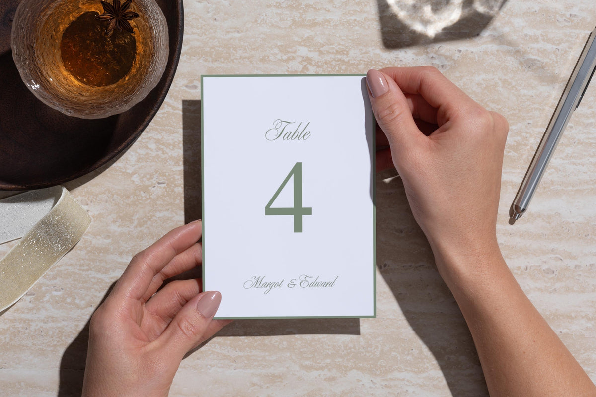 Sage green wedding table number template viewed from above, held over a textured neutral table beside a drink and cutlery, with refined serif typography for understated old money wedding reception décor.
