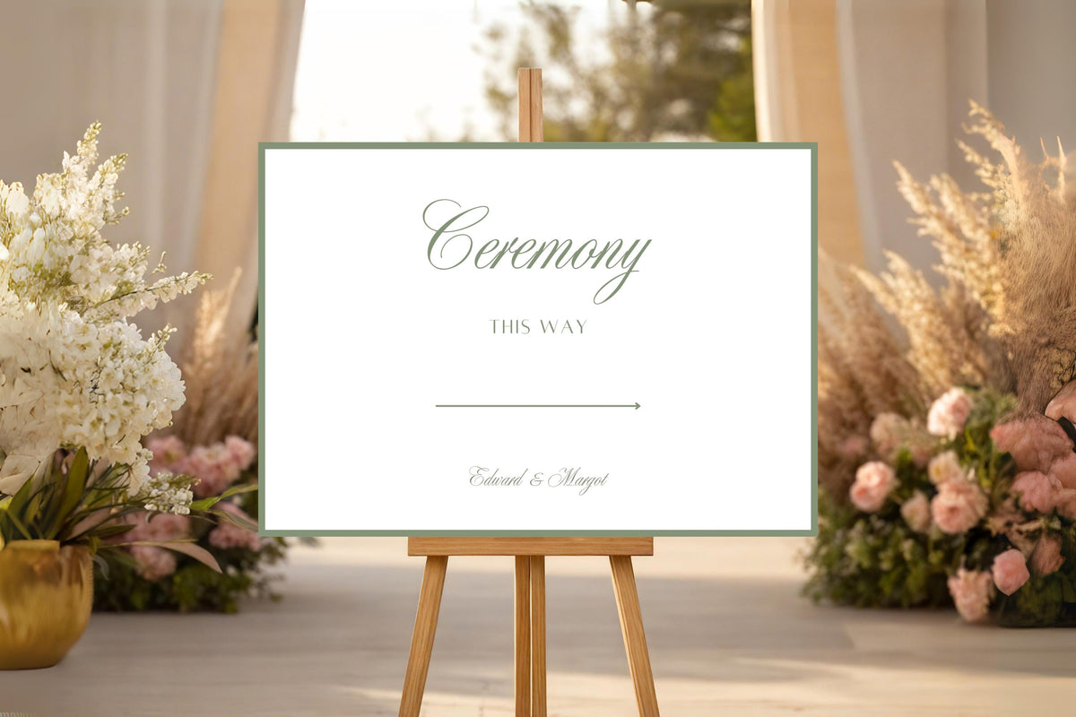 Minimal white wedding ceremony sign with a sage green border, displayed on an easel among blush and cream flowers at a romantic wedding venue.