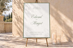 Ivory wedding welcome sign with a sage green border and romantic script names, displayed on a wooden easel beside glass pillar candles against a sunlit stone wall for an elegant outdoor wedding entrance.