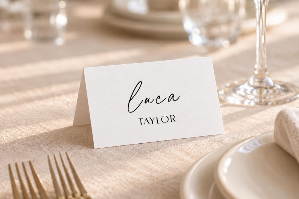 Minimalist folded wedding place card on a neutral linen reception table beside gold cutlery and white crockery, featuring a guest name in elegant black script for a personalised wedding table setting.