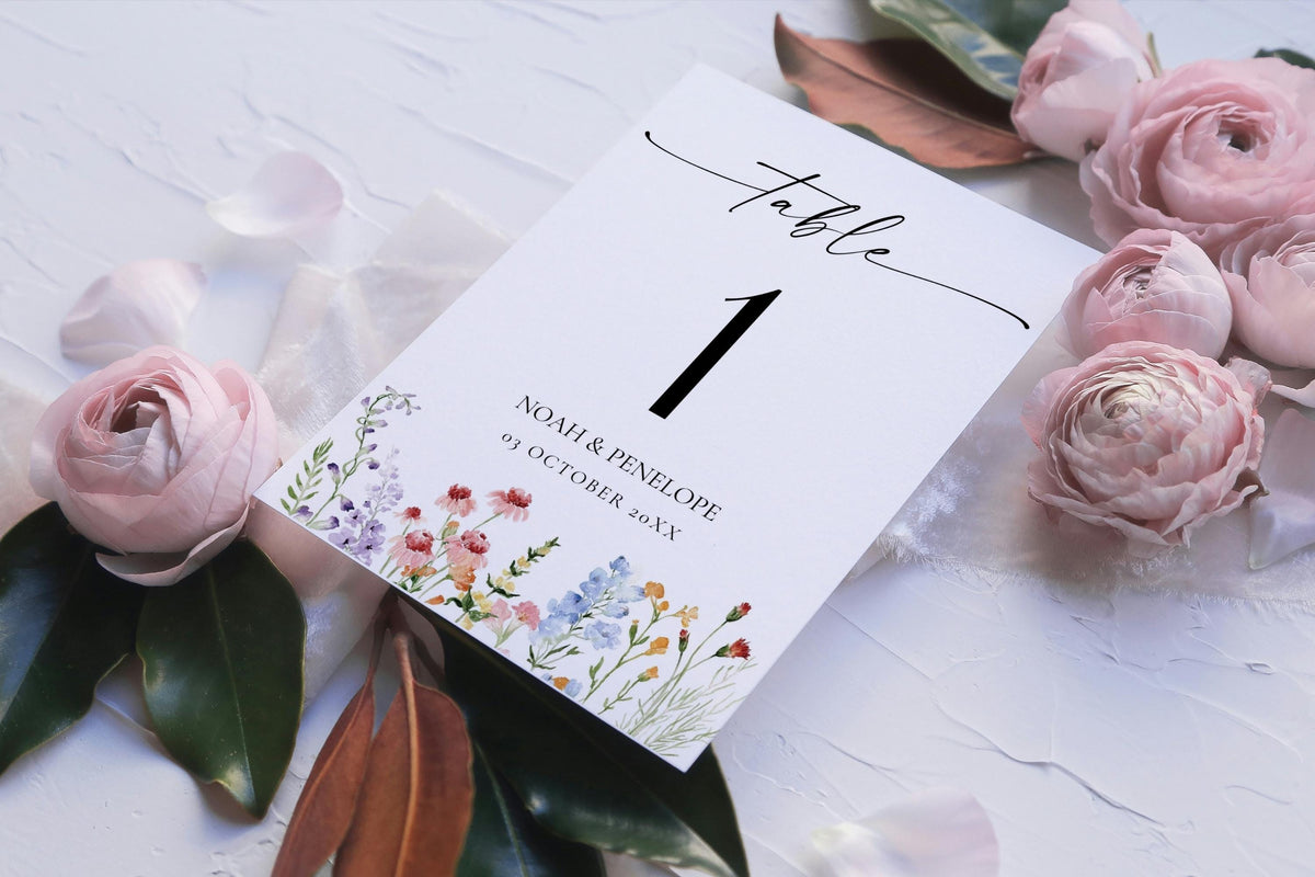 Flat lay of floral wedding table number card with watercolor wildflowers, editable printable wedding stationery template for rustic boho reception decor.