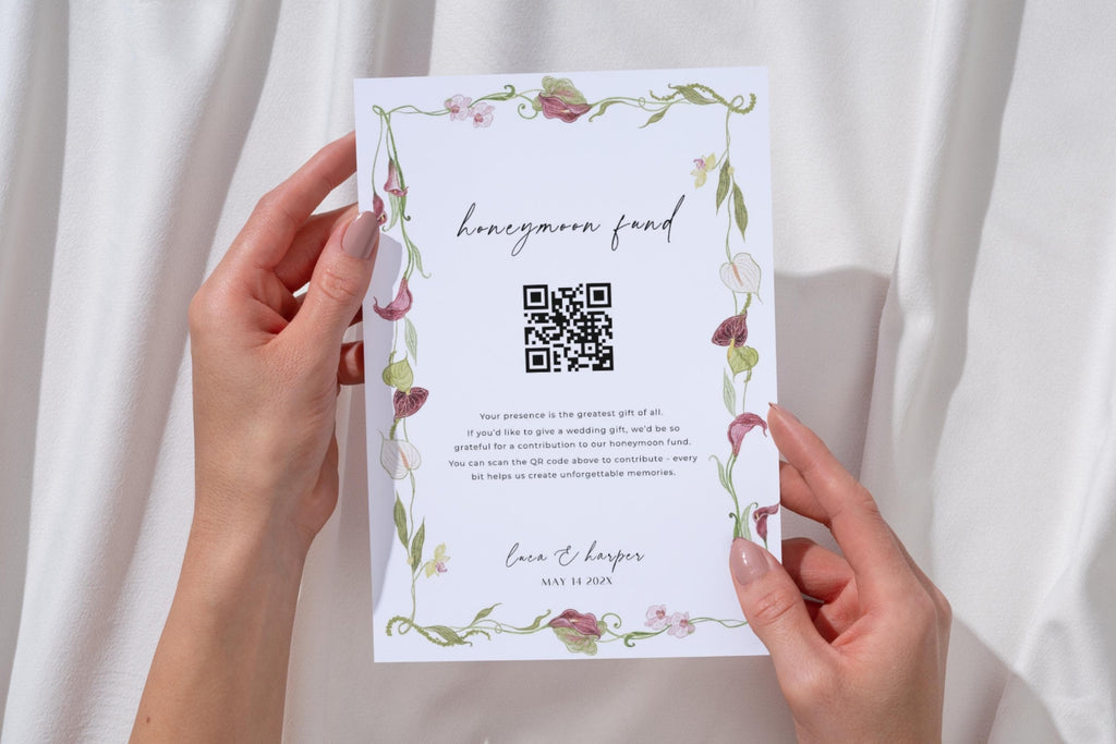 Editable honeymoon fund wedding sign with QR code, held against white fabric and decorated with pink flowers and green foliage for a coordinated wedding gift table display.