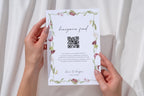 Editable honeymoon fund wedding sign with QR code, held against white fabric and decorated with pink flowers and green foliage for a coordinated wedding gift table display.