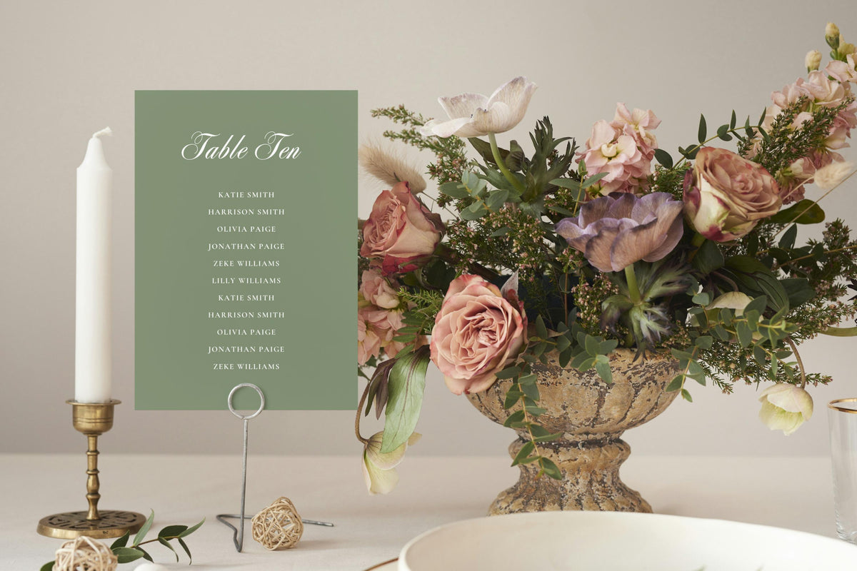Sage green individual wedding seating chart sign displayed beside a floral centrepiece and taper candle, featuring white script lettering for a romantic garden wedding reception.