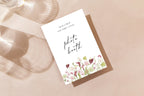 Printable floral wedding photo booth sign on a neutral tabletop, featuring black handwritten lettering and a pink flower border for a romantic wedding reception photo station.