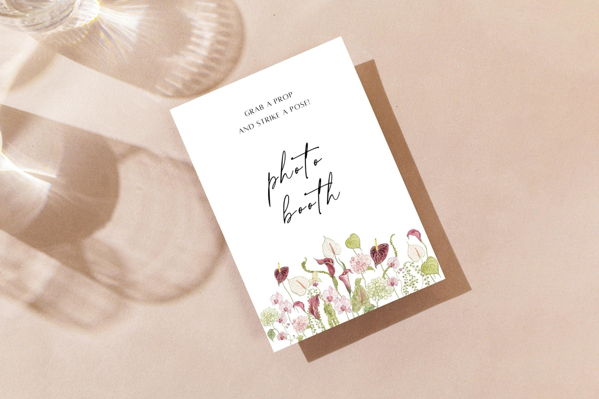 Printable floral wedding photo booth sign on a neutral tabletop, featuring black handwritten lettering and a pink flower border for a romantic wedding reception photo station.