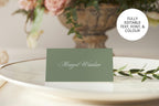 Solid sage green folded wedding place card with delicate white script lettering, displayed on a white plate beside greenery and flowers for an elegant garden wedding reception.
