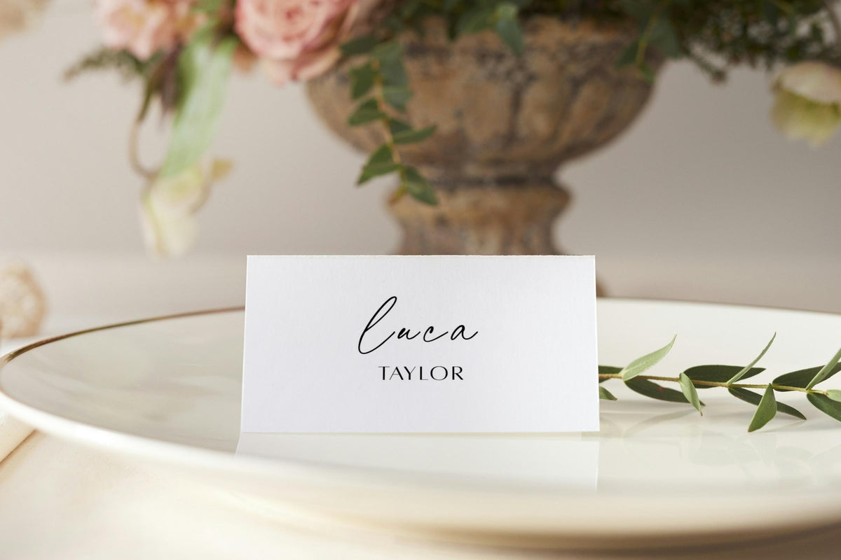 Minimalist folded wedding place card with elegant script name, styled beside white tableware and soft pink floral wedding décor.