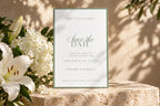 Sage green wedding save the date card displayed on natural stone beside white lilies and hydrangeas. Classic bordered stationery template for a romantic garden wedding or elegant destination celebration.