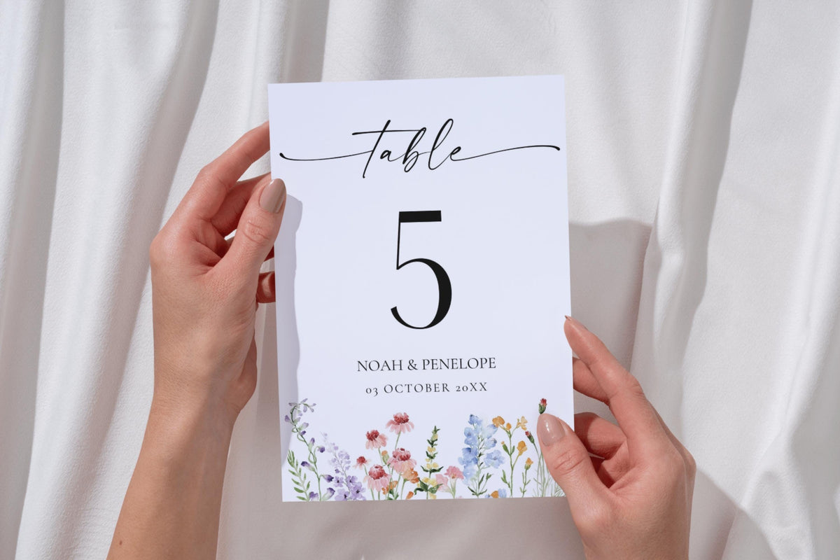 Hands holding wildflower wedding table number card, editable floral Canva template for DIY printable wedding reception table decor.