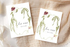 Matching his and her vows booklet templates with elegant script lettering, green trailing botanical borders and burgundy flowers. Personalised wedding ceremony vow books displayed on soft beige fabric in natural light.