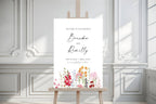 Romantic floral wedding welcome sign shown on an easel against white decorative panelling, with editable names and a bright, whimsical flower design.
