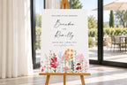 Editable wedding welcome sign displayed on a wooden easel beside bright glass doors, featuring a romantic multicolour floral design and elegant script names, Brandon and Kennedy.