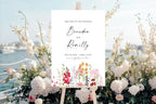 Colourful floral wedding welcome sign displayed beside the sea, styled with pale blue water, white flowers and personalised names for a romantic coastal wedding.