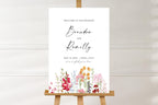 Customisable wedding welcome sign mockup against a soft white wall, featuring colourful floral illustrations and elegant script names for a romantic wedding entrance.