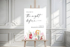 Close-up of a colourful floral rehearsal dinner welcome sign on an easel against a white panelled wall.