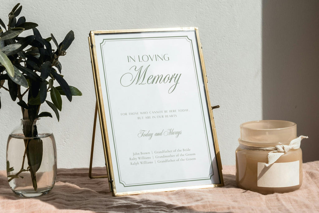 In loving memory wedding sign with sage green lettering and a fine border in a gold frame, displayed beside greenery and candles for a thoughtful wedding memorial table.