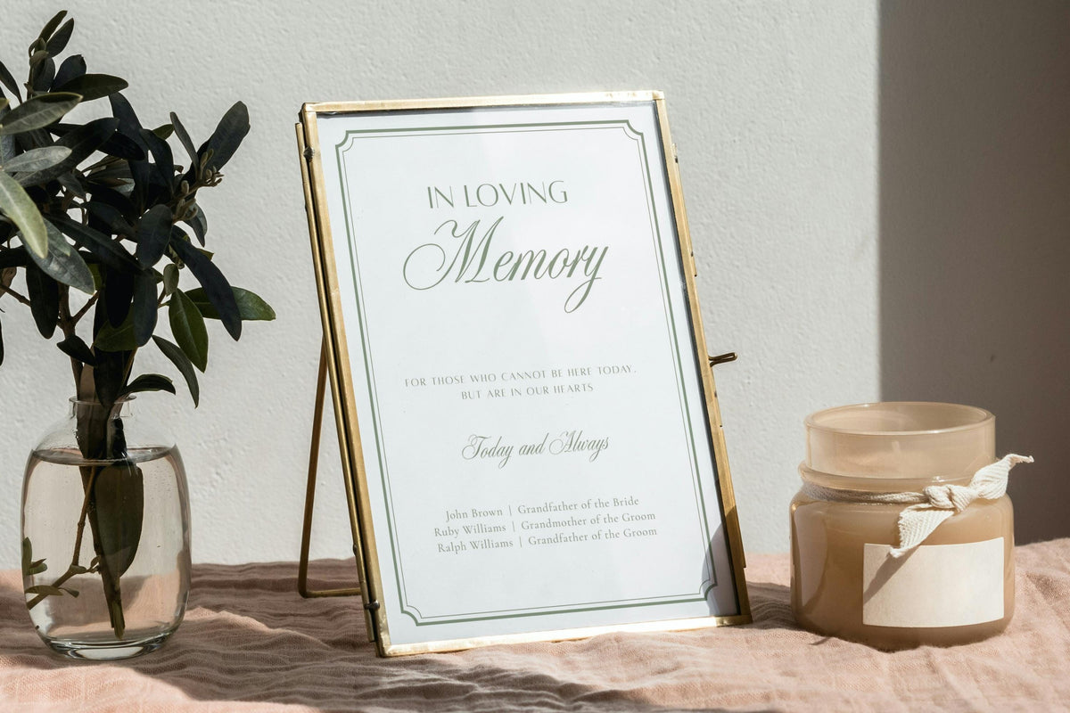 In loving memory wedding sign with sage green lettering and a fine border in a gold frame, displayed beside greenery and candles for a thoughtful wedding memorial table.