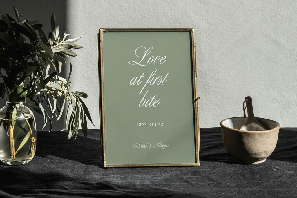 Sage green Love at First Bite wedding dessert table sign with white calligraphy in a gold frame, styled on dark linen beside greenery and a small serving bowl.
