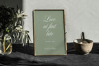 Sage green Love at First Bite wedding dessert table sign with white calligraphy in a gold frame, styled on dark linen beside greenery and a small serving bowl.