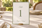 Sage green and ivory wedding table number card with a large serif numeral, delicate script and classic border, displayed in a small brass holder on a sunlit reception table with elegant place settings.