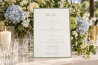 Wedding bar menu sign with sage green typography on a white background, styled with blue and white hydrangeas, glassware and candles for a classic wedding reception drinks table.