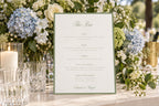 Elegant ivory wedding bar menu sign with a sage green border and script heading, displayed beside crystal glassware, candlelight and blue and white flowers for a romantic garden wedding drinks station.