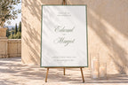 Personalised sage green wedding welcome sign with elegant script names and a fine border, displayed on a wooden easel beside glass candle holders in a sunlit stone courtyard.