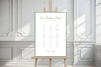 Minimalist sage green wedding seating plan displayed on an easel against white panelled walls with natural sunlight and shadows. Old money wedding table chart featuring elegant script, a green border and organised guest lists.