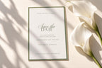 Sage green bordered save the date card with elegant script and classic serif lettering on ivory cardstock, styled beside white calla lilies and soft fabric with natural shadows for an old money wedding.