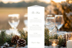 Classic trifold wedding menu displayed on an outdoor reception table with evergreen foliage, pine cones, candles and wine glasses, providing inspiration for elegant winter wedding table stationery.