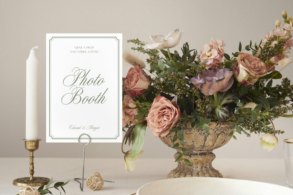 Elegant wedding photo booth sign with sage green calligraphy and a fine decorative border, displayed beside pastel flowers and a tall taper candle on a reception table.