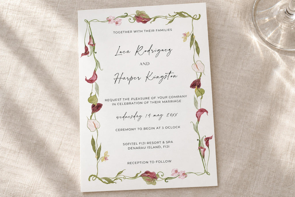 Romantic botanical wedding invitation template with delicate pink flowers and winding green stems framing elegant script names, photographed on ivory linen beside a glass for a soft garden wedding look.
