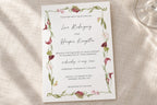 Romantic botanical wedding invitation template with delicate pink flowers and winding green stems framing elegant script names, photographed on ivory linen beside a glass for a soft garden wedding look.