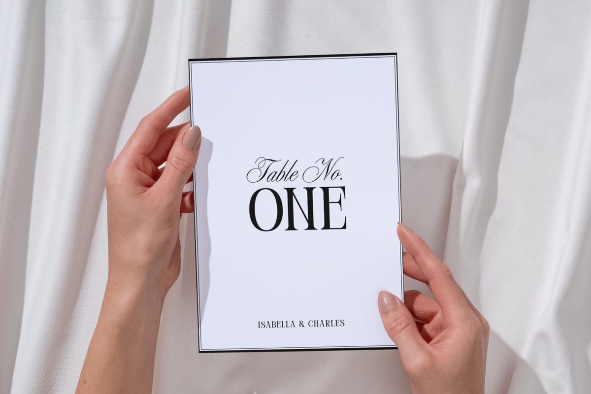 Hands holding minimalist wedding table number print, editable black serif table number template for classic modern wedding décor, printable table number signage for reception tables.