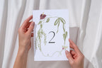 Bride holding an editable floral wedding table number two, featuring whimsical wildflowers and a delicate botanical border.