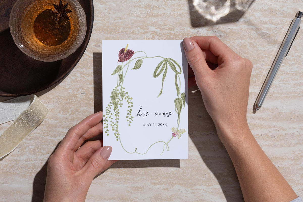 Hands holding a personalised her vows wedding booklet on a stone tabletop beside a pen and drink. Green botanical and burgundy floral cover design for a romantic garden wedding vow book and ceremony keepsake.