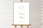 White and sage green “The Night Before” rehearsal dinner sign featuring a thin green border, classic serif lettering and script names. Minimalist wedding welcome sign displayed on a wooden easel against a neutral wall.