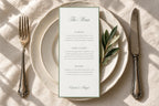 Sage green wedding menu template with elegant script heading and thin green border, styled on ivory plates with an olive sprig, textured linen and vintage cutlery for an old money wedding reception.
