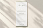 Minimalist sage green wedding menu card on a plain ivory background with soft sunlight and shadows, featuring a script heading, refined serif text and slim border for timeless old money wedding stationery.