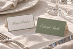 Folded sage green and ivory wedding place cards with contrasting script guest names, styled on a white reception table beside silver cutlery and crystal glassware for elegant old money wedding table decor.