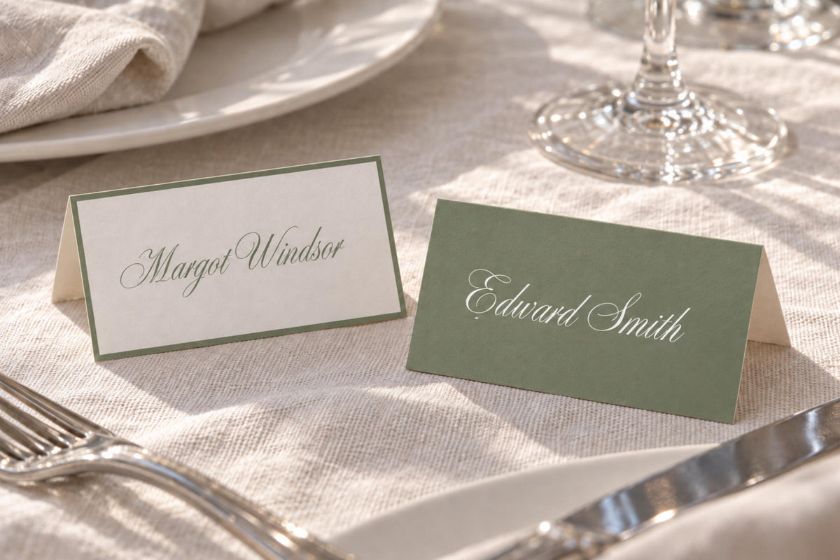 Folded sage green and ivory wedding place cards with contrasting script guest names, styled on a white reception table beside silver cutlery and crystal glassware for elegant old money wedding table decor.